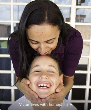 Victor, who lives with hemophilia A, is shown seated with his eyes closed and smiling while his mother stands behind him, holding his face with her hands 